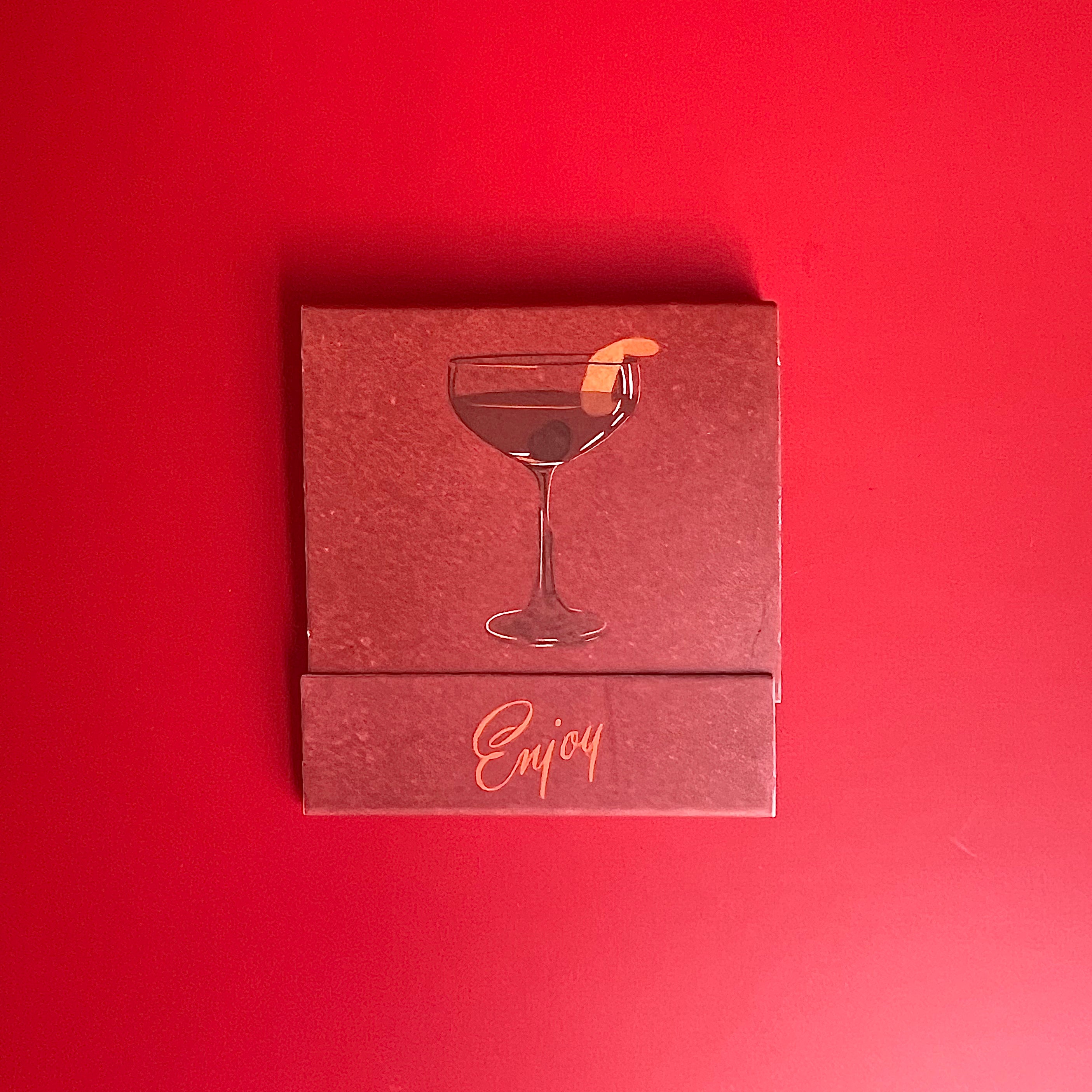 Manhattan cocktail matchbook featuring a red background with a drawing of a manhattan in a glass and the word, “Enjoy!”