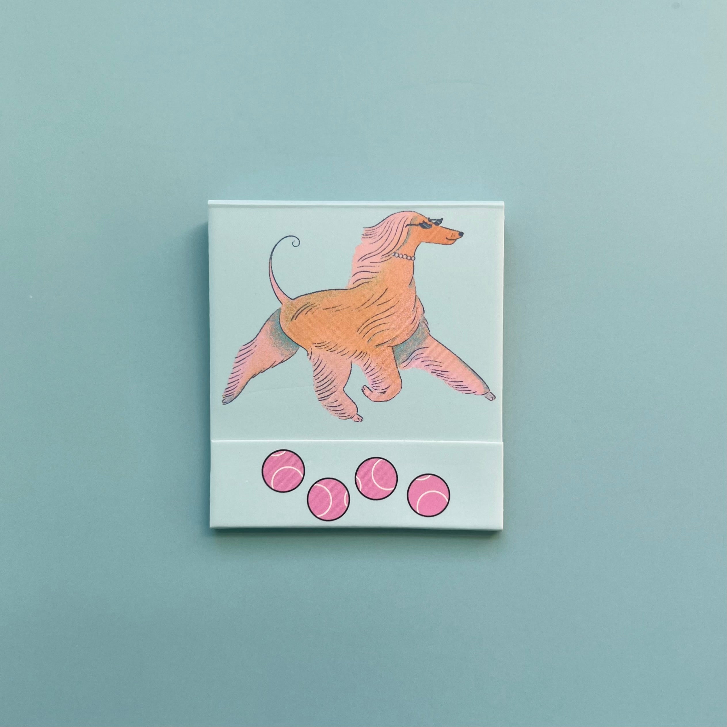 Front of dog matchbook featuring an illustration of an Afghan Hound with tennis balls on the bottom