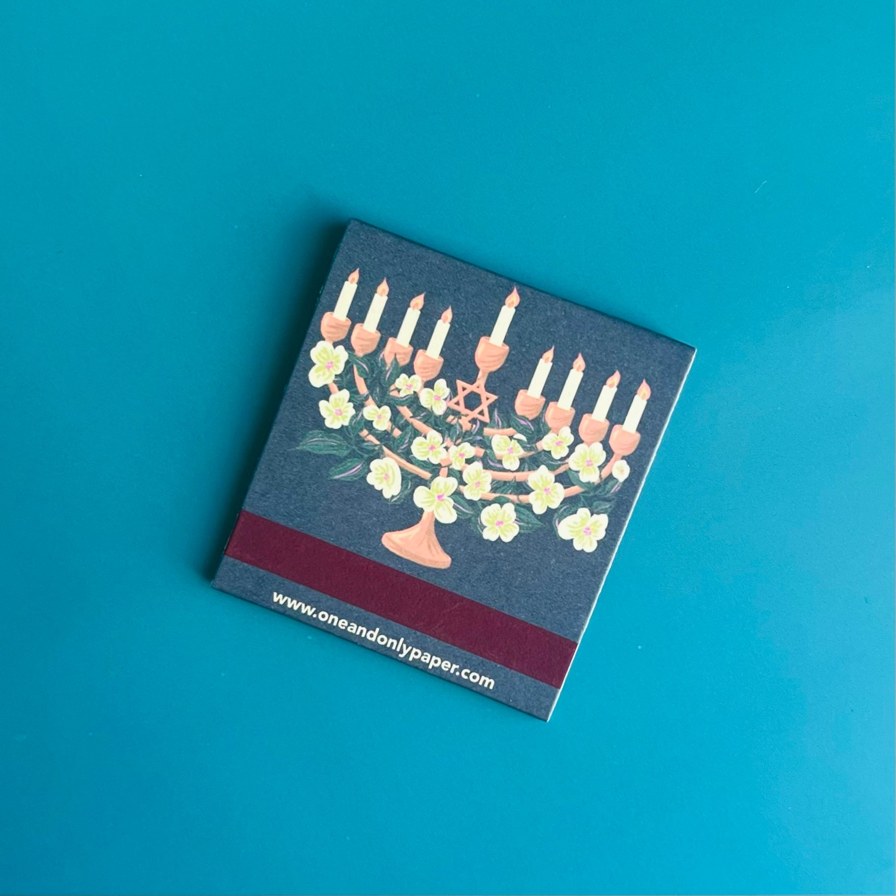 Back of menorah matchbook depicting an illustration of the memorial with florals interwoven and the Star of David with match-striking bar on the bottom