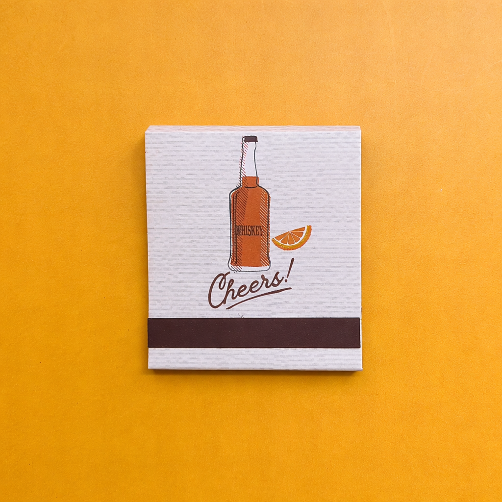 Back of cocktail matchbook featuring the drawing of a whiskey bottle, an orange slice, and the word, “Cheers!”
